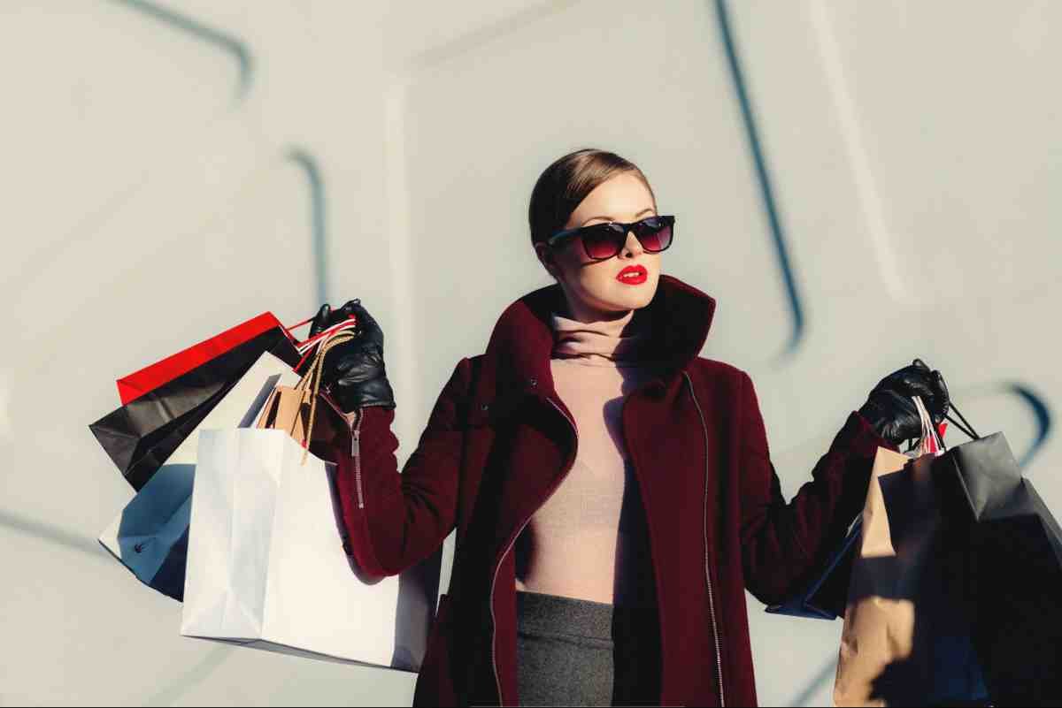 Fashion dropshipping how to start: woman in burgundy coat and sunglasses holding colorful shopping bags outdoors
